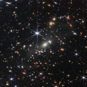 Distant galaxies appear as bright glowing spots in this Webb telescope image, with some smeared by gravitational lensing; foreground stars appear bright with six-pointed diffraction spikes, owing to the shape of Webb's mirrors